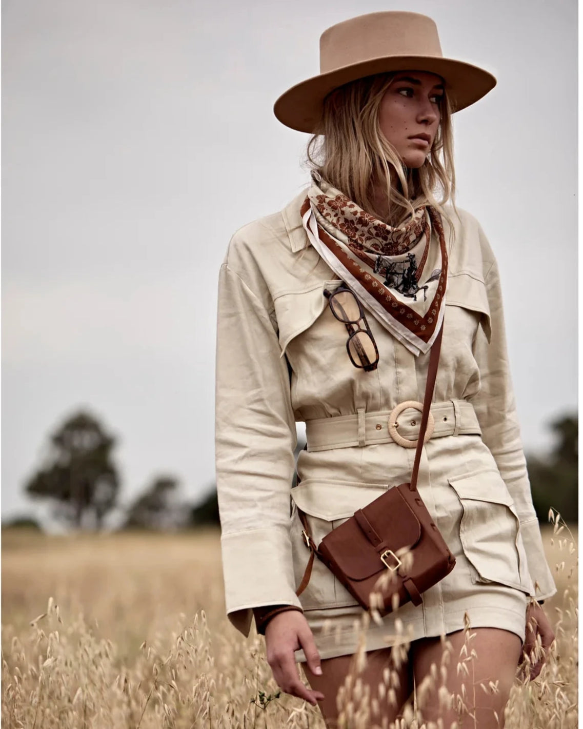 Woman in a beige trench coat and hat standing in a field, holding a brown bag.
