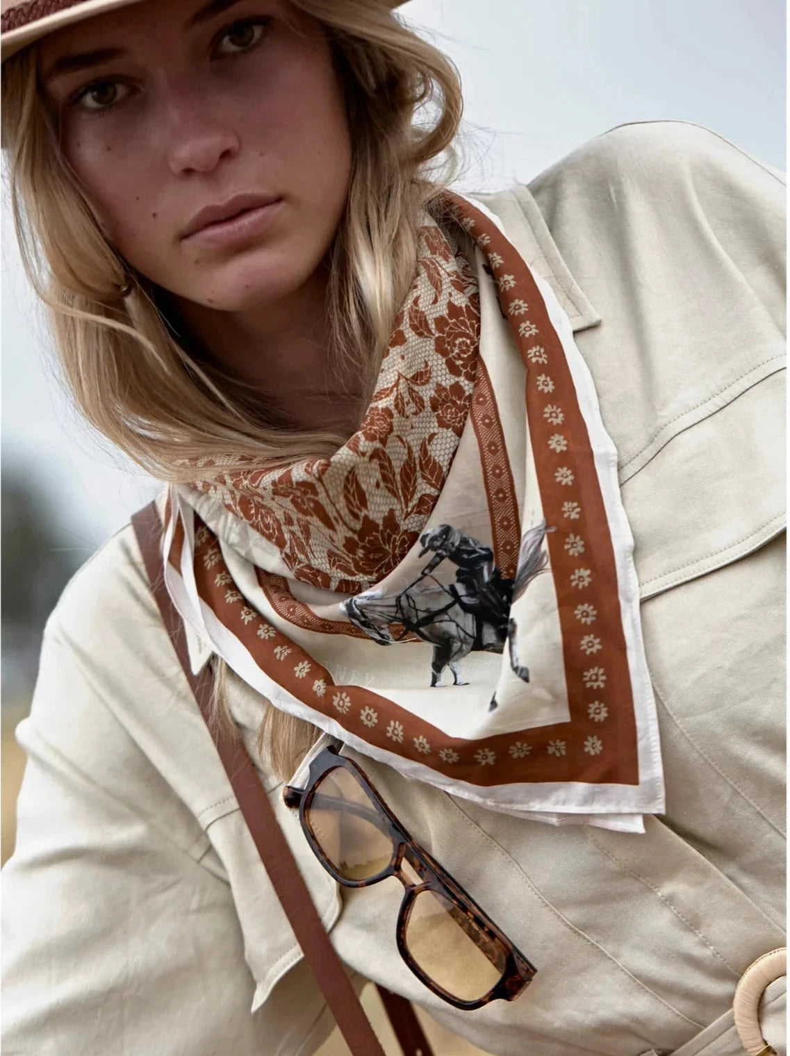 Woman wearing a patterned scarf with a horse design, holding glasses, and standing outdoors.