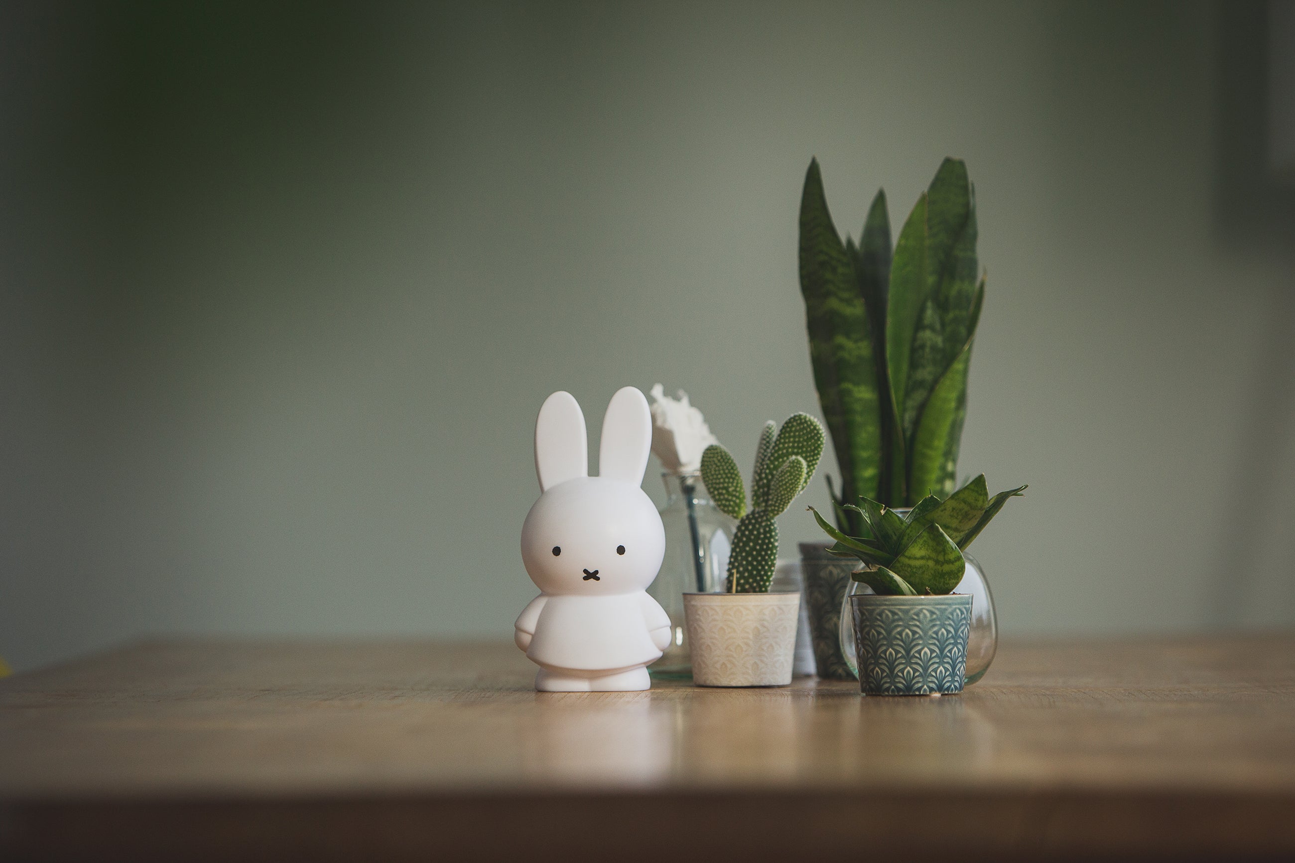 A small white rabbit figurine stands on a wooden table beside potted green plants in a softly blurred indoor setting.