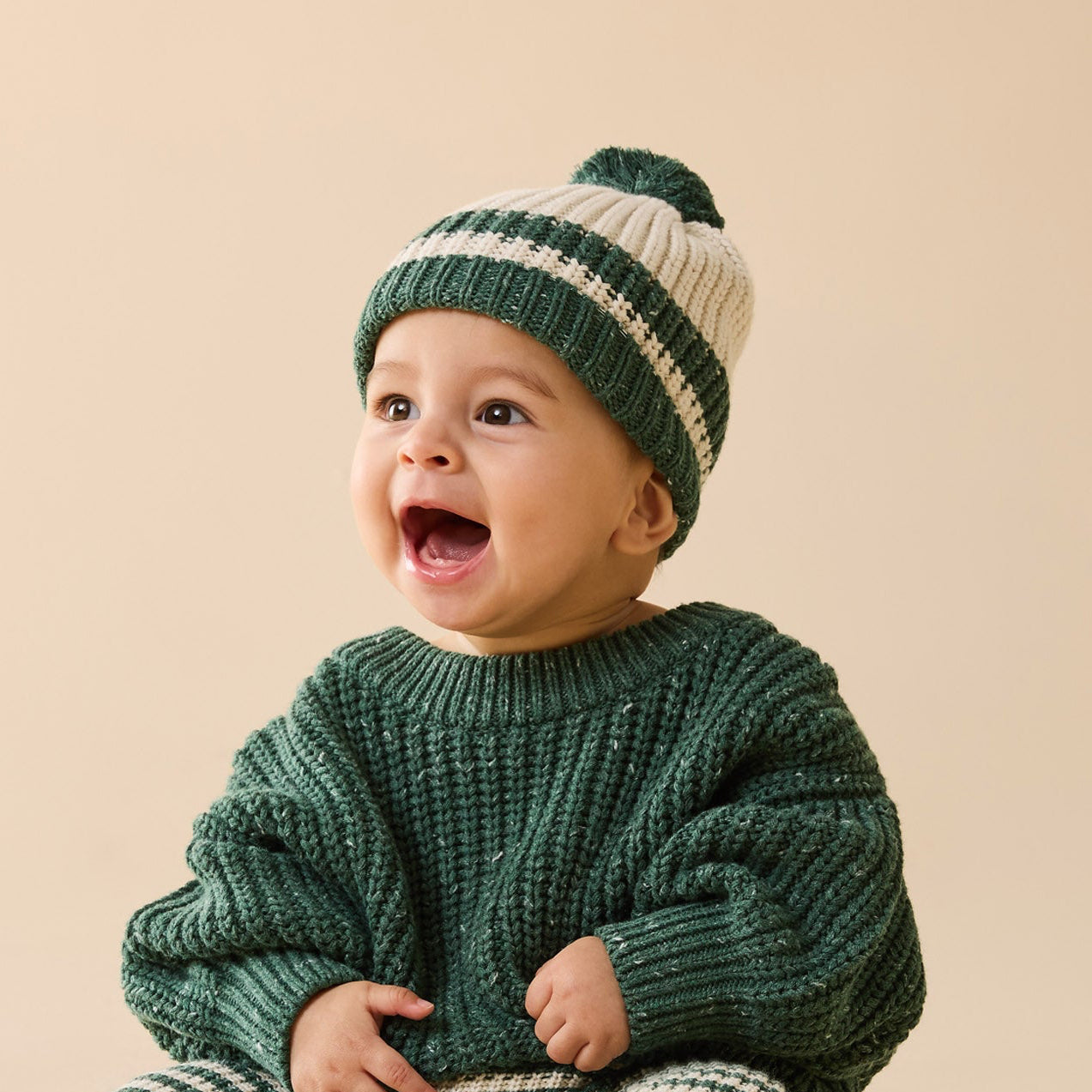 Baby wearing a green knitted outfit with a matching hat on a beige background