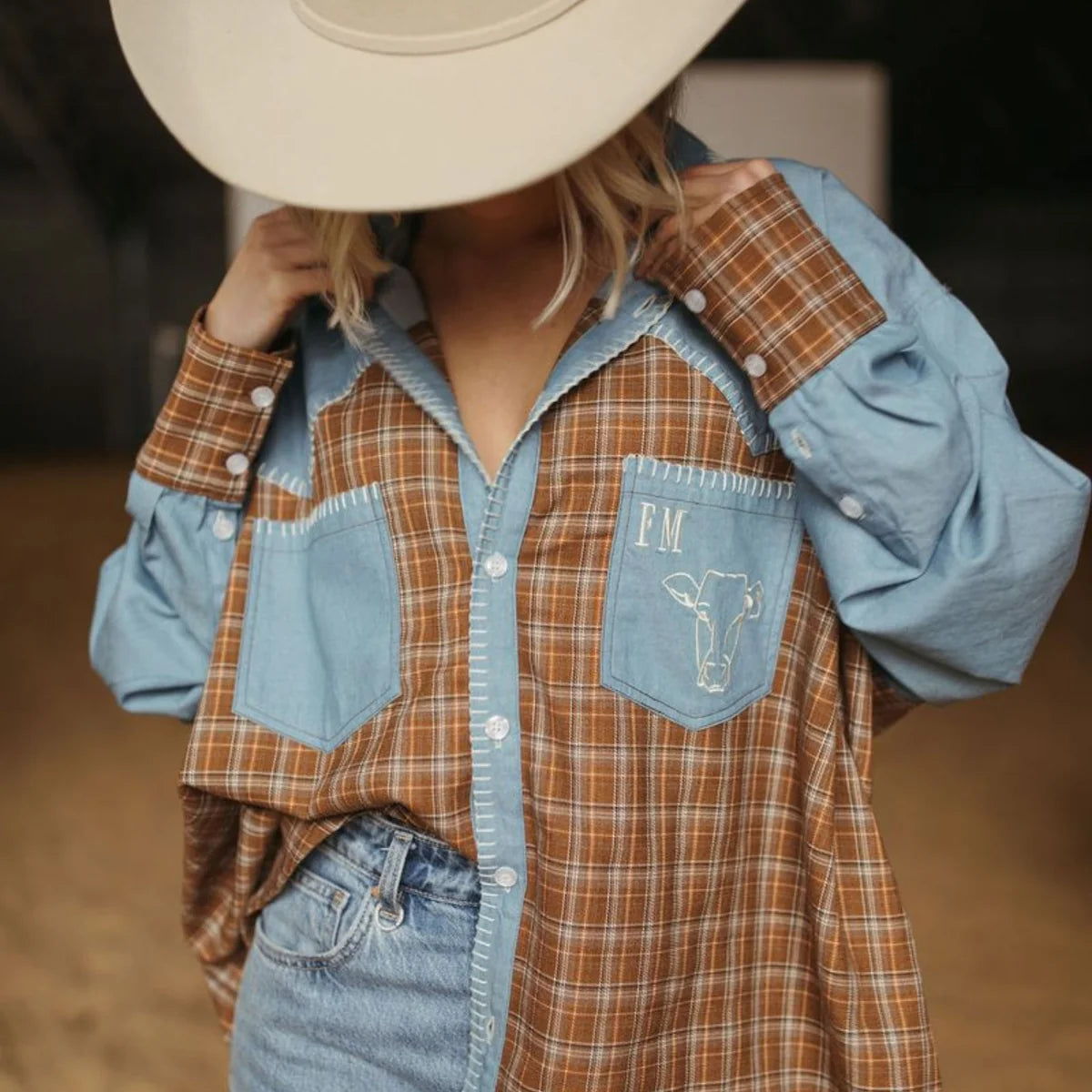 A person wearing a color-block western-style plaid shirt with light blue and brown sections, a matching cowboy hat, and denim jeans in a rustic indoor setting.