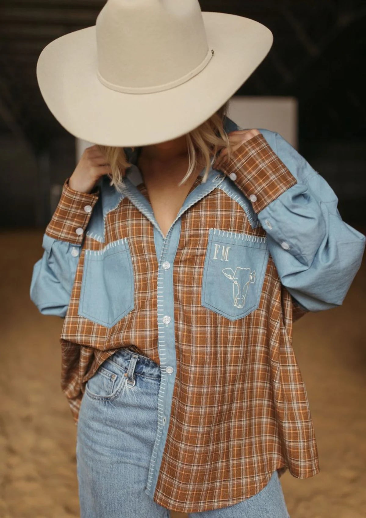 A person wearing a color-block western-style plaid shirt with light blue and brown sections, a matching cowboy hat, and denim jeans in a rustic indoor setting.
