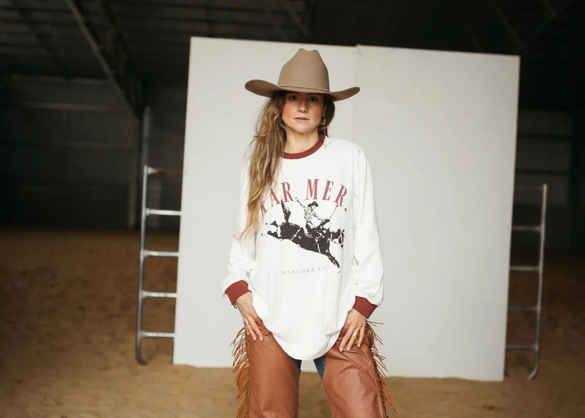 A woman modeling a white long-sleeve graphic sweatshirt with red trim and fringe, paired with brown leather chaps and a cowboy hat, standing in a studio with a white backdrop and sandy floor.