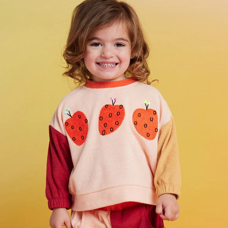 A young child wearing a color-blocked knit sweater with three strawberry appliques on the front, set against a solid yellow background.
