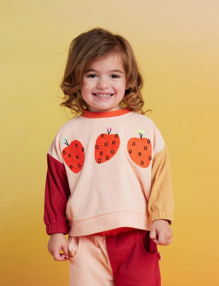 A young child wearing a color-blocked knit sweater with three strawberry appliques on the front, set against a solid yellow background.